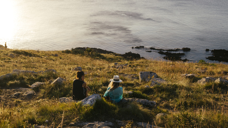 Two people sitting on grassy bluff overlooking ocean at sunset