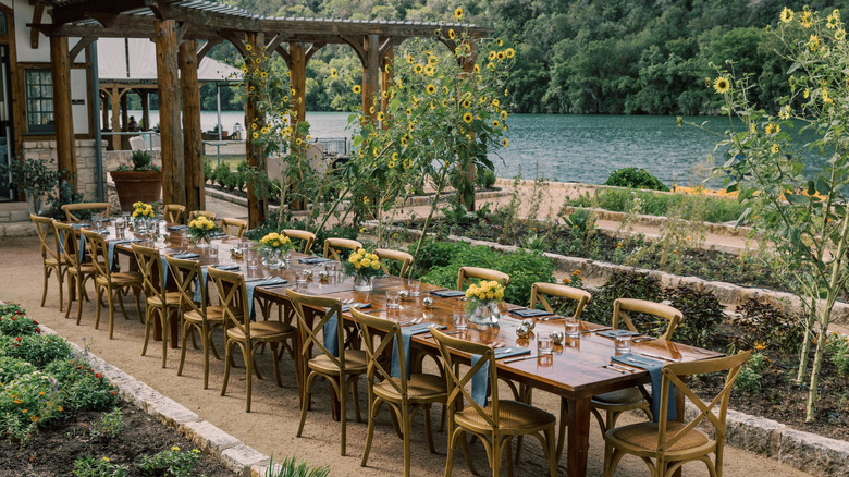 A long table with chairs by a pergola, greenery, and a riverfront