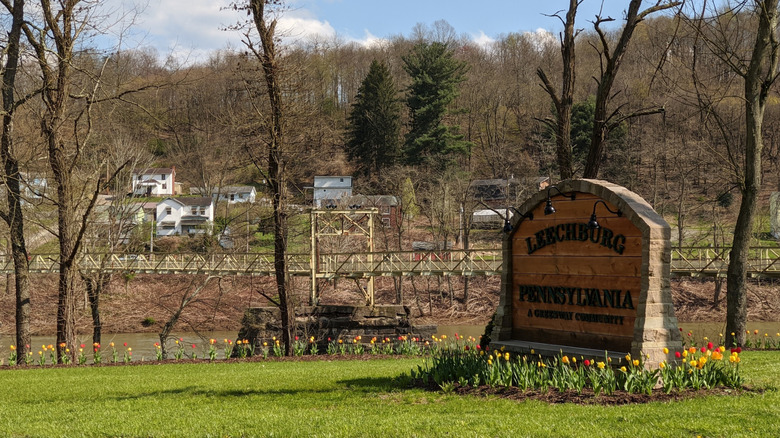 Wooden sign reading "Leechburg Pennsylvania" ringed by yellow and red tulips, with a river in the background crossed by a yellow footbridge and white homes and leafless trees on the far shore, under a cloudy blue sky