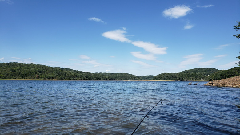 Blue lake with a fishing pole in the foreground and treed hills in the background, under a cloudy blue sky
