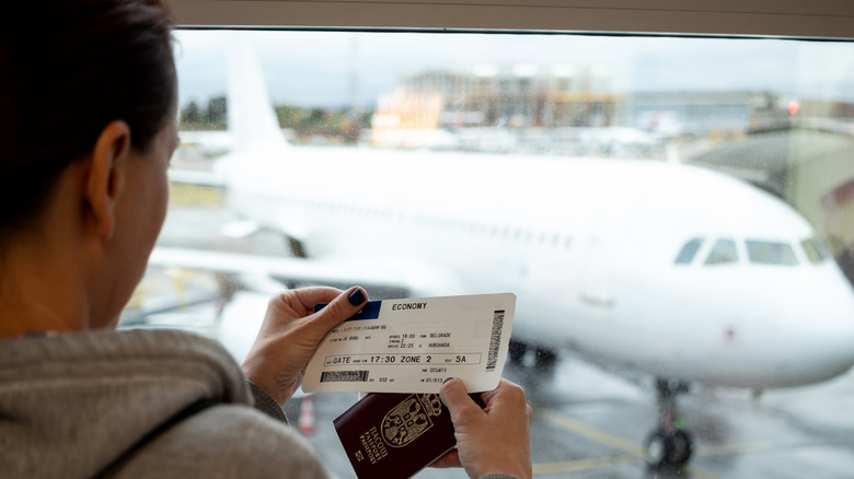 Traveler holding a boarding pass and passport while looking at a plane through the airport window