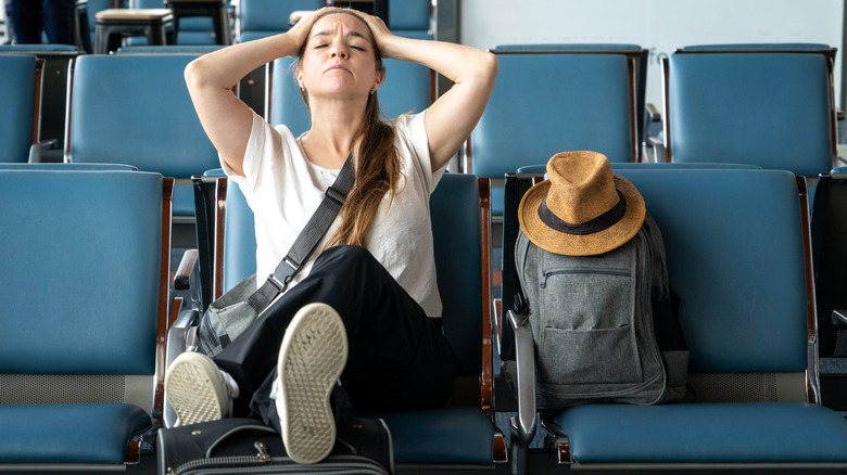 Tired traveler sitting in airport chairs, holding head with hands beside luggage and hat