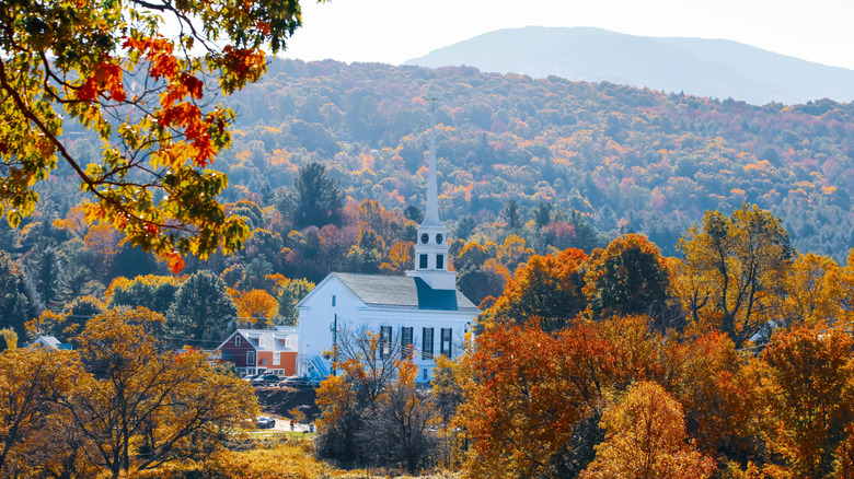 A steepled white church is surrounded by hills and autumnal leaves in Vermont on a sunny day