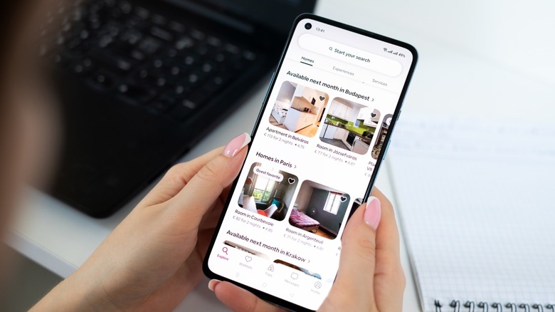A woman looks at Airbnb listings on a phone with a laptop and notebook in the background
