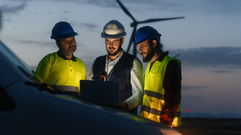 An electrical engineer and industry workers discuss a project at a renewable energy station, with a windmill in the background, over a laptop at night