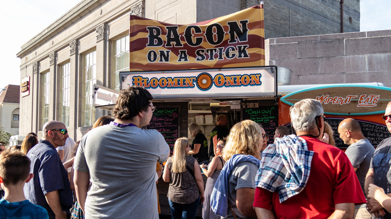 People gathered outside in front of a food truck selling bacon on a stick and bloomin' onion.