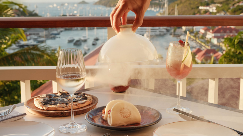 A hand lifting a glass dome filled with smoke off of a plate revealing bao buns on a dining table overlooking the Caribbean Sea at Beefbar restaurant at Fouquet's Saint-Barth