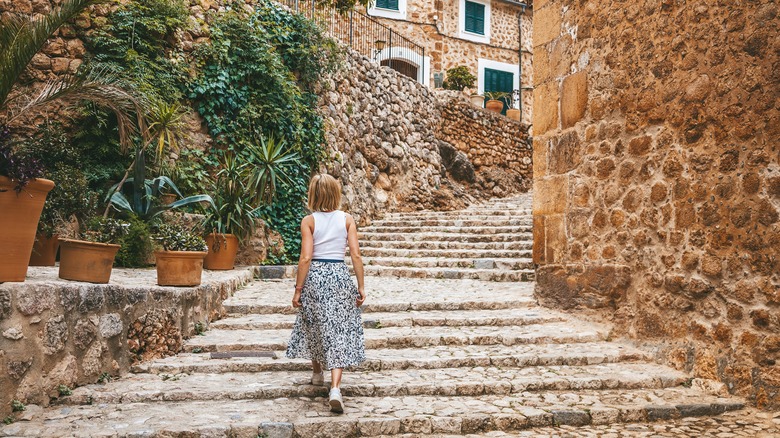 A woman in a white top and patterned skirt walks up cobblestone steps in Fornalutx