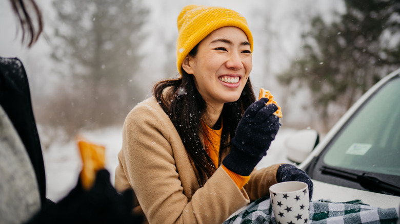 Woman in a yellow beanie holding a hot drink on top of her car