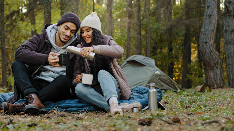 Young couple sits in a forest smiling and pouring coffee from a thermos into a cup
