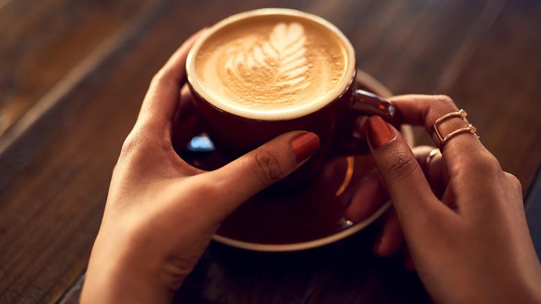 Close-up of a woman's hands holding a mug of coffee, designed with latte art, on a wooden table
