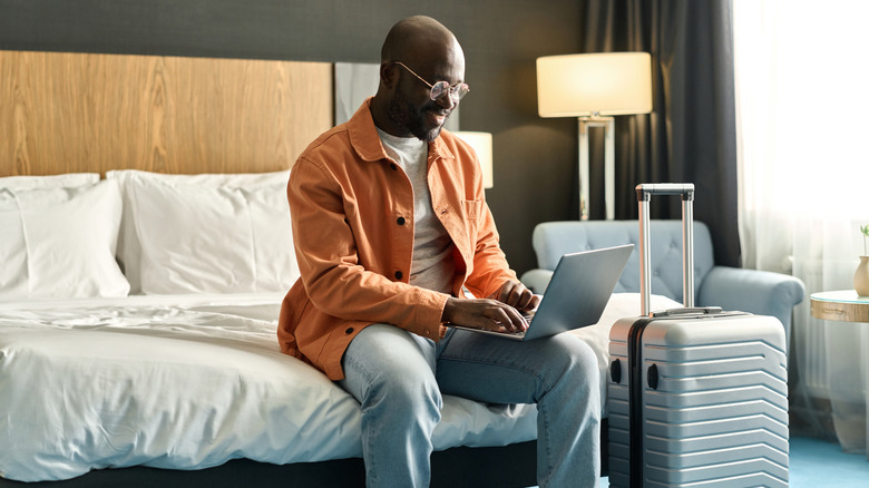 Man sitting on hotel bed using laptop with luggage beside him