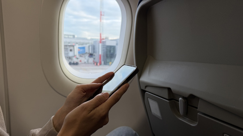A pair of hands holding a cell phone next to the window seat in a plane