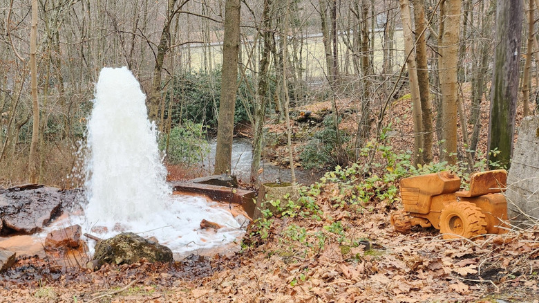 Geyser of water on the left side of the frame, with an orange-stained tractor on the right, surrounded by leafless trees with a creek and yellow house in the background