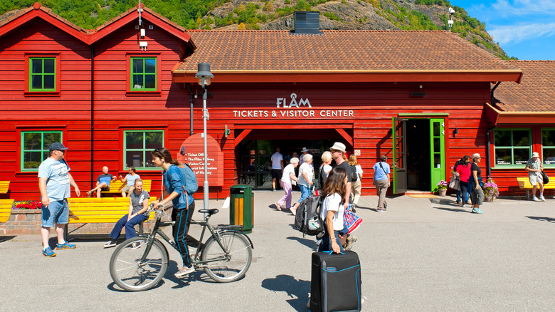 Travelers with bicycles and suicases pass in front of the red Flåm train station