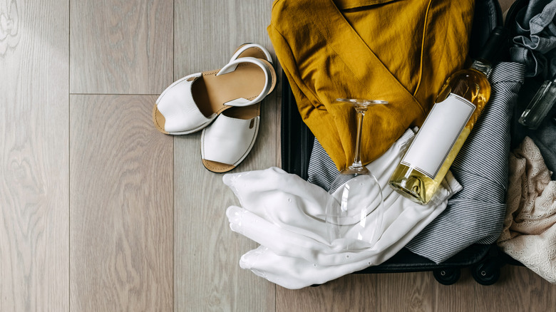Open suitcase full of summer clothes, a wine glass and a bottle of white wine.