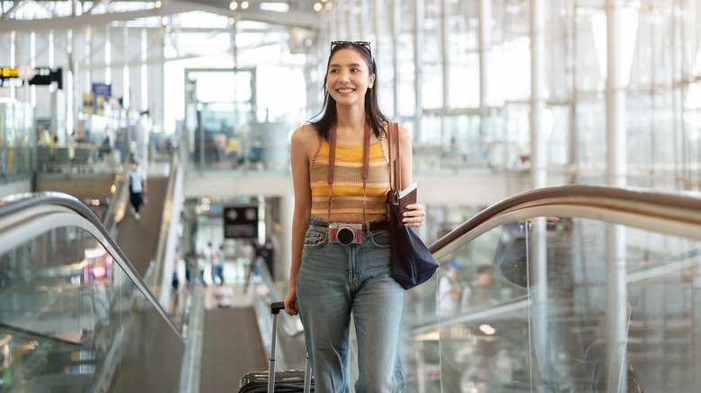 Beautiful young smiling Asian woman with camera and luggage ascends escalator in unidentified airport terminal