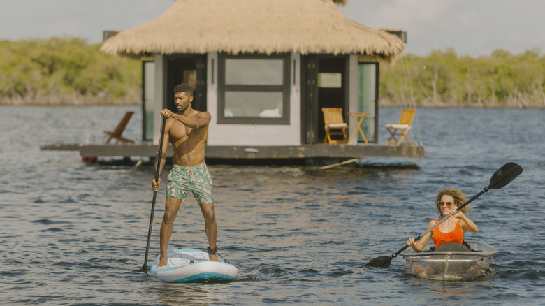 a man on a paddleboard and woman on a kayak paddle away from an overwater bungalow
