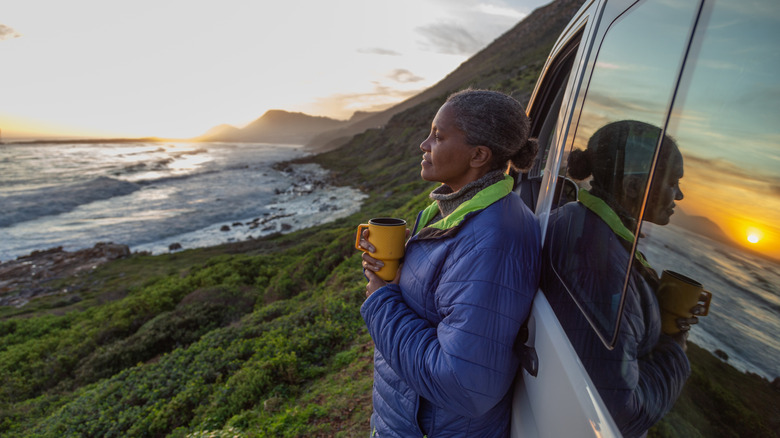 A retired woman holds a cup of coffee and enjoys a stunning view leaning against her RV.