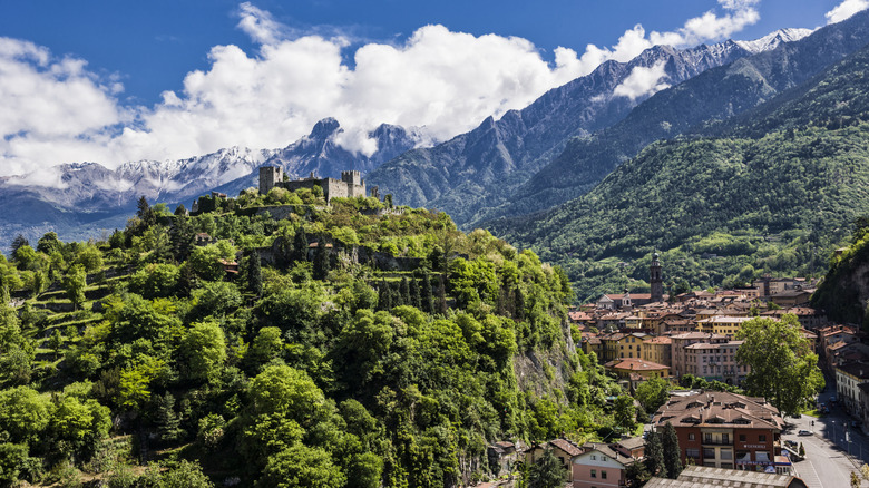 A castle sits on a bluff below cloud-covered mountains in the Alps