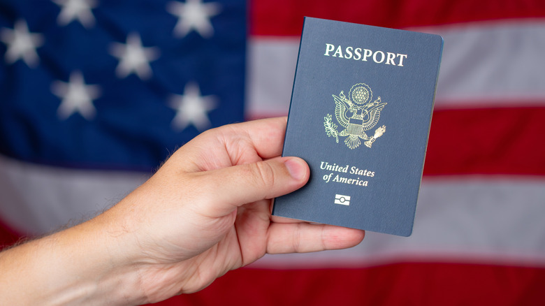 A hand holding a US passport in front of an American flag.