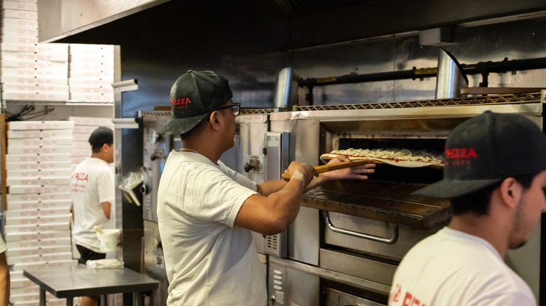 Joe's Pizza employee putting a pizza in the oven, with many pizza boxes in the background