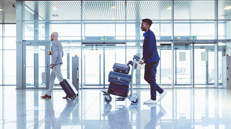Man pushing a luggage cart with three suitcases in an airport while another passenger rolls a suitcase past