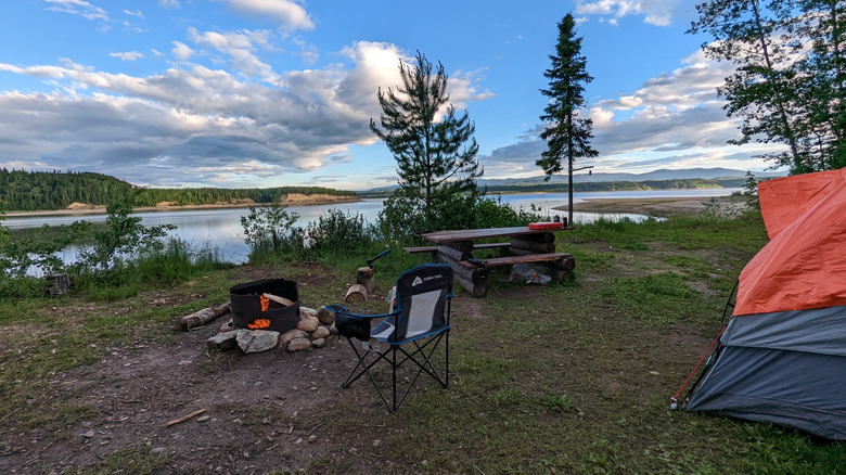 View of a camping set-up with a tent, chair, picnic table, and fire pit on the shore of a lake