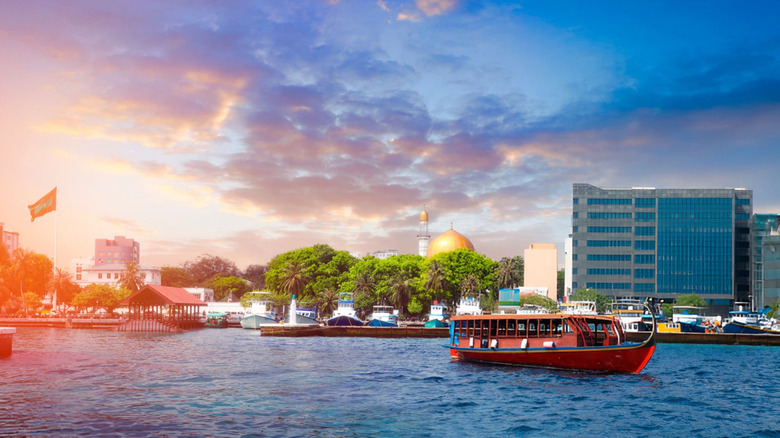 Skyline of the city of Malé, Maldives, viewed from the water, as the sun sets over buildings, boats, and a mosque