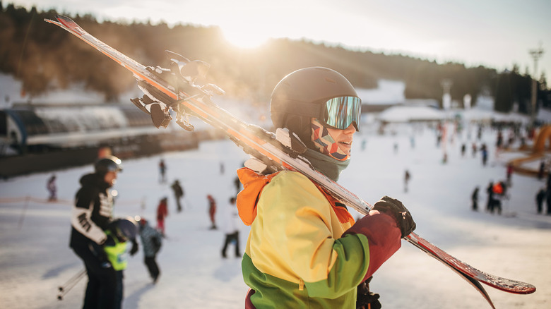 A skier holding skis as the sun sets behind a snowy hill