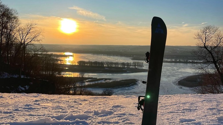 A snowboard sticking vertically out of the snow on a hill overlooking the Mississippi at sunset