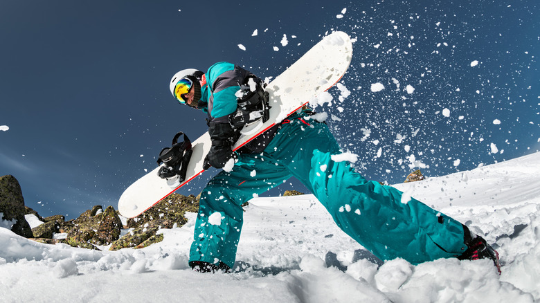 A snowboarder walking in the sun with their board and snow falling off of it