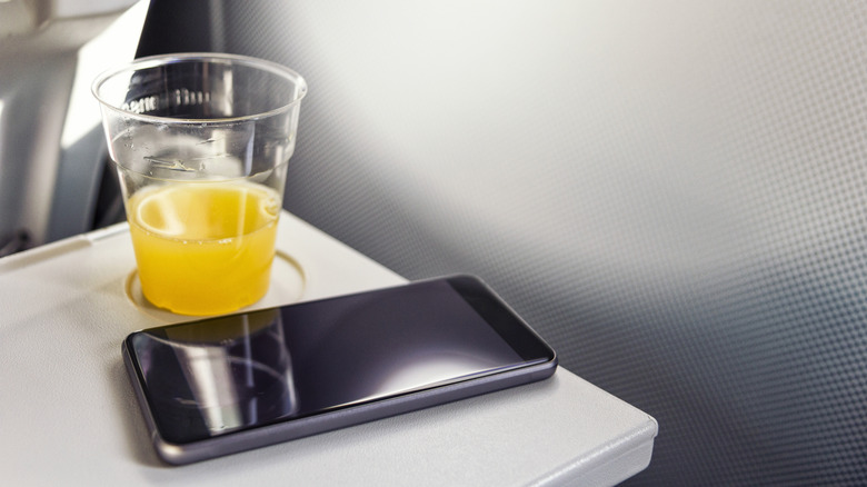 A cup of juice sat next to a phone on a plane seat tray.