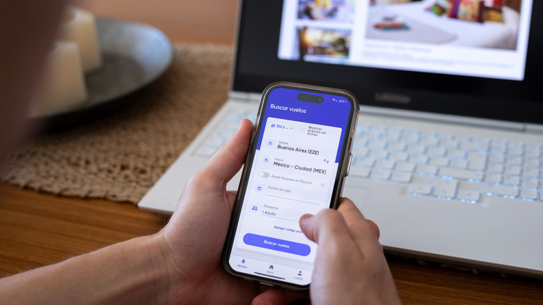 Close-up of hands searching for flights from Buenos Aires to Mexico using mobile phone with laptop and dining table in background