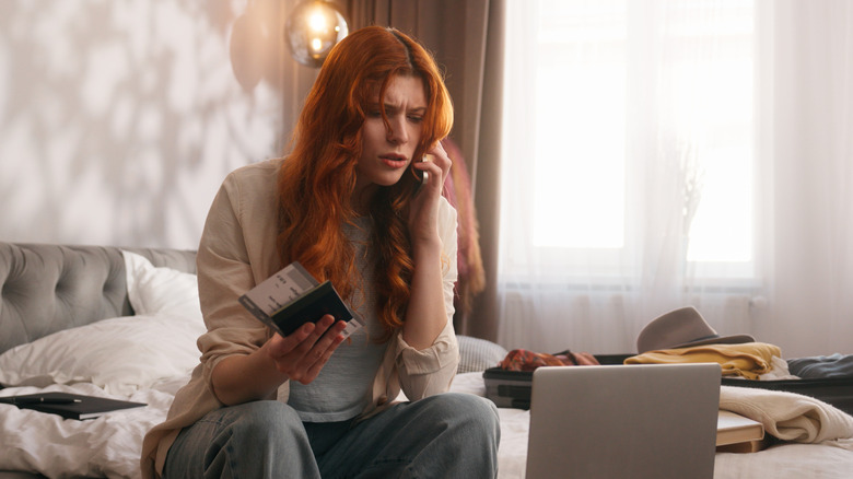 Frustrated redhead woman sitting on her bed next to a laptop while talking on cell phone, with passport in one hand, and a suitcase behind her, presumably talking to an airline
