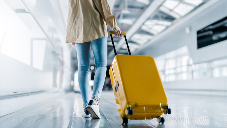 International airport terminal, close-up of woman in skinny jeans and a tan shirt pulling bright yellow suitcase