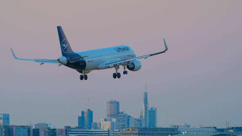 Lufthansa plane descending toward an airport with a backdrop of a modern city skyline at dusk.