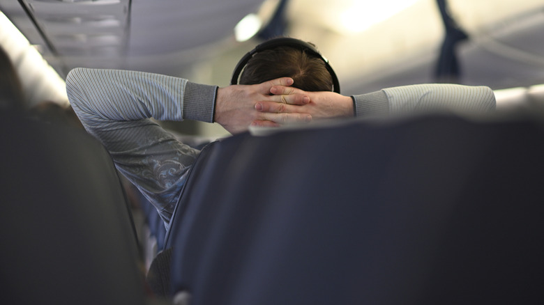 Back view of male passenger relaxing in a comfortable airline seat with hands behind his head and headphones on