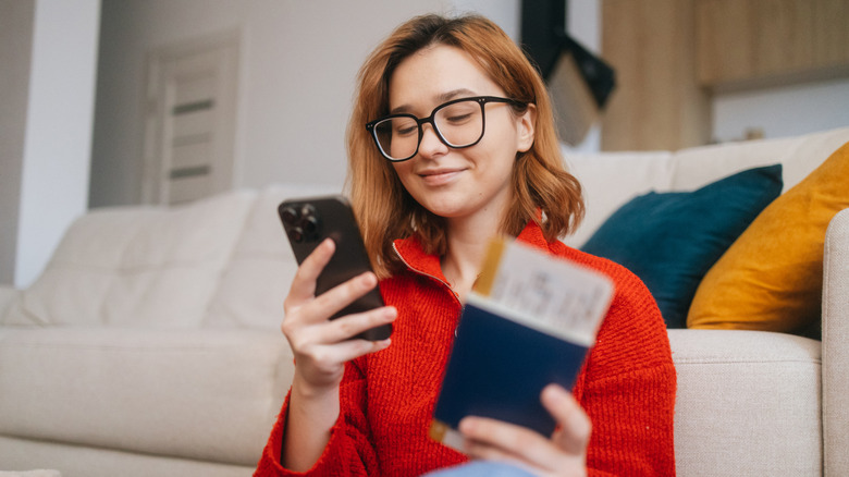 A cheerful woman uses her smartphone to make an online flight booking while holding her passport in the other hand