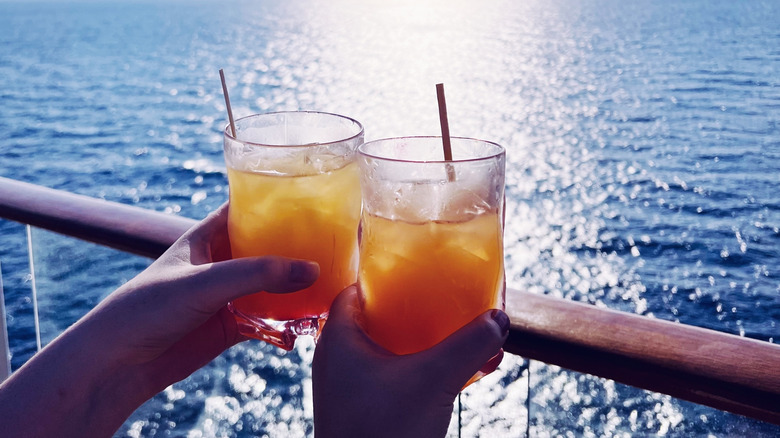 Close-up of couple's hands clinking glasses with tropical cocktails on a cruise ship deck with sun-dappled water in the background