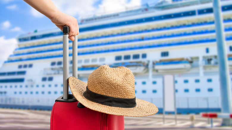 Close up of a hand holding a red suitcase with a rattan hat on top of it, with blurred cruise ship in the background