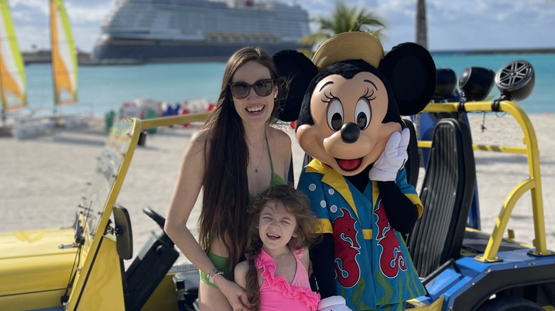Brunette mother and daughter smile and pose with Minnie Mouse on Disney's Castaway Cay private island with Disney Wish ship blurred in the background