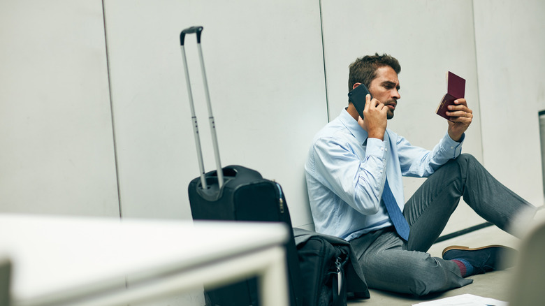 Young man sitting on airport floor talking on the phone with passport in hand