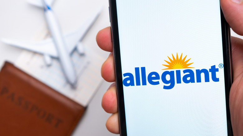 Allegiant airlines company app or logo displayed on a mobile phone with passport, boarding pass, and plane on the background