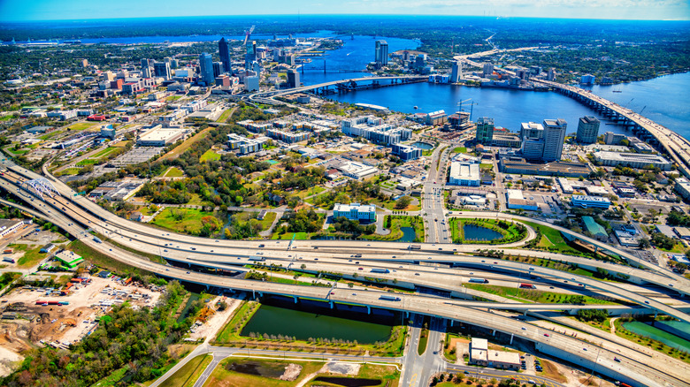 A network of highways, including I-95, criss-crosses like veins in Jacksonville, Florida, with trees, a river, limited sky, and buildings