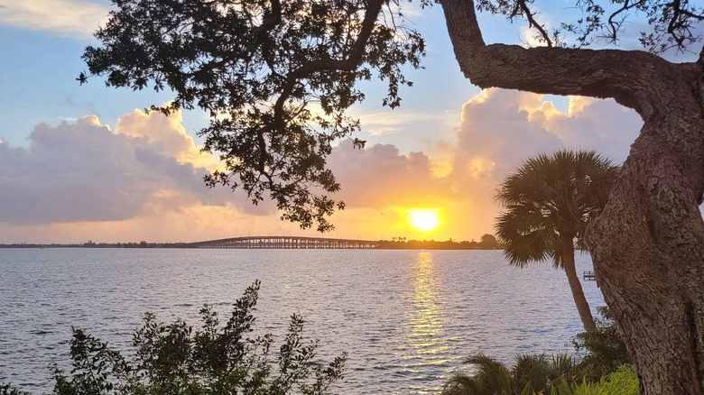 Distant view of a bridge along U.S. 192 with trees in the foreground, a large lake, a sunset, and evening-lit clouds