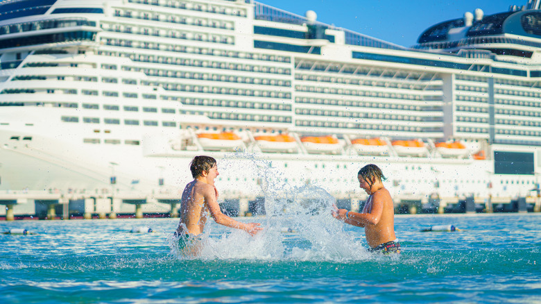 Two boys splashing each other in the water near a cruise ship