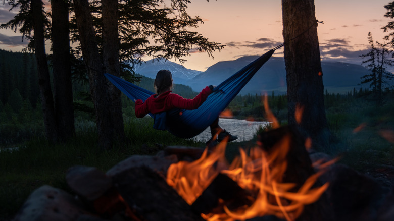Camper relaxes in a hammock staring at mountains with a campfire in the background