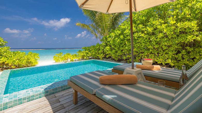Blue and white striped pool chairs beside a small rectangular pool, overlooking a beach under a blue sky
