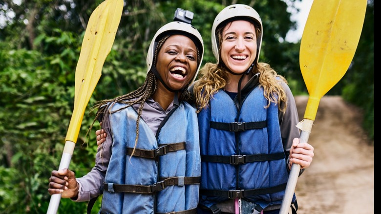 Two smiling young women hold paddles and wear helmets and lifejackets